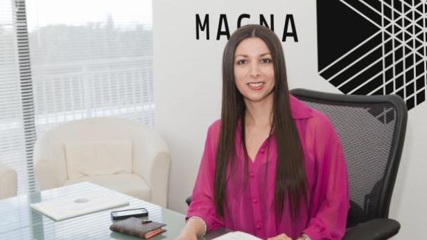 Eine Frau sitzt an einem Schreibtisch vor dem Magna-Logo.