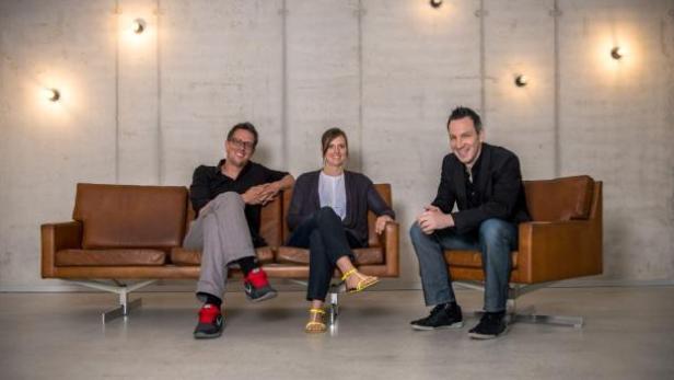 Drei Personen sitzen auf einer Ledercouch und Sesseln vor einer Betonwand mit Lampen.