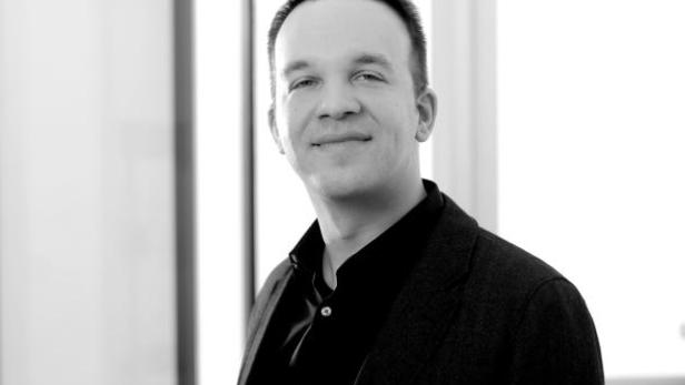 Ein Mann in einem dunklen Anzug und Hemd lächelt in die Kamera.