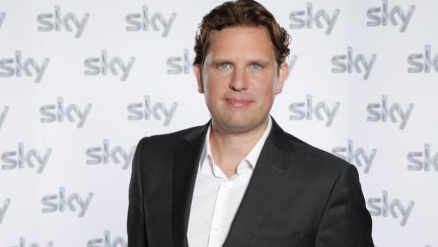 Ein Mann im Anzug steht vor einer Wand mit dem Sky-Logo.