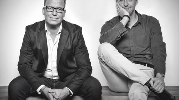 Zwei Männer sitzen auf einer Bank vor einer weißen Wand.