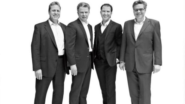 Vier Männer in Anzügen stehen nebeneinander vor einem weißen Hintergrund.