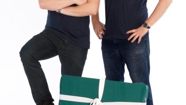 Zwei Männer mit Simplewish-T-Shirts stehen hinter mehreren bunten Geschenken.