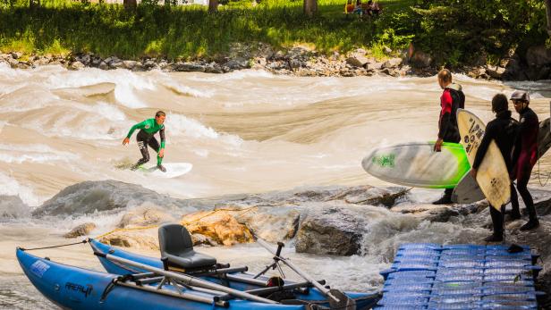 Ein Surfer reitet auf einer Welle in einem Fluss, während andere mit Surfbrettern am Ufer stehen.