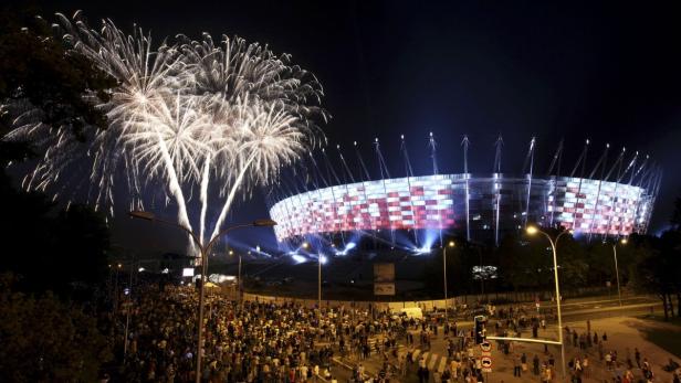 Feuerwerk erleuchtet den Himmel über einem beleuchteten Stadion und einer Menschenmenge.