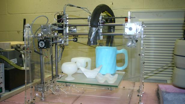 Ein 3D-Drucker fertigt verschiedene Objekte, darunter eine Tasse und dekorative Formen.