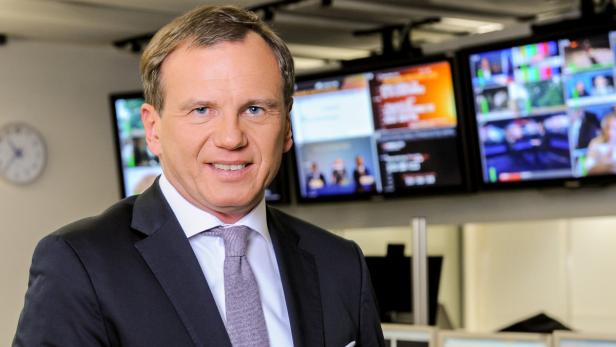 Ein Mann im Anzug lächelt vor mehreren Monitoren.