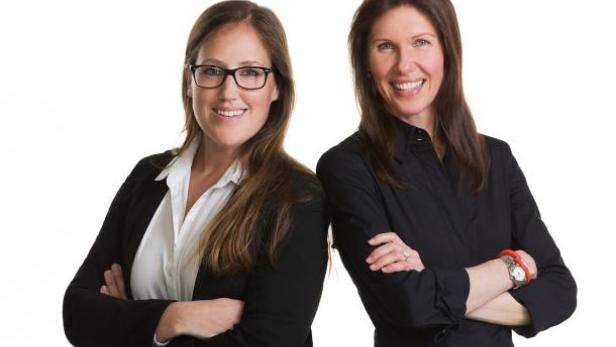 Zwei Frauen stehen mit verschränkten Armen vor einem weißen Hintergrund.