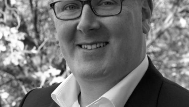 Ein lächelnder Mann mit Brille und Anzug vor einem unscharfen Hintergrund.