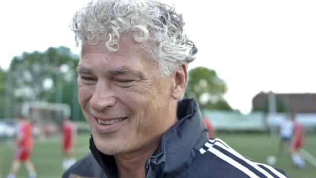 Ein Mann mit grauem, lockigem Haar lächelt auf einem Fußballplatz.