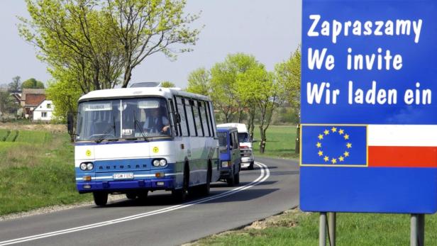 Ein Bus fährt an einem Schild vorbei, das auf Polnisch, Englisch und Deutsch „Wir laden ein“ sagt.