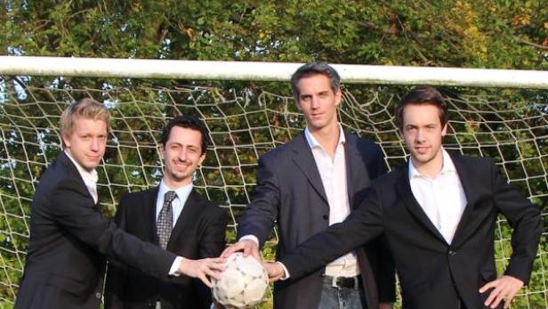 Vier Männer in Anzügen halten einen Fußball vor einem Fußballtor.