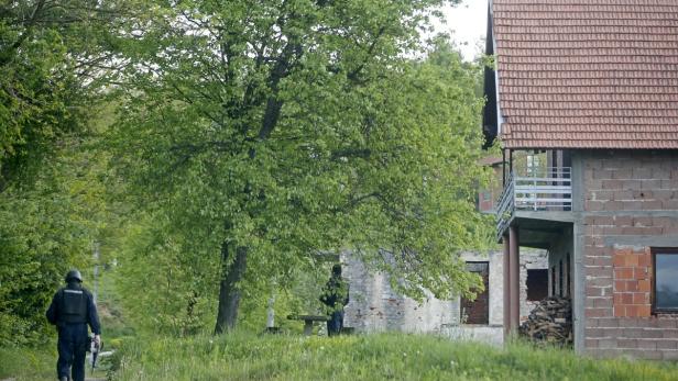 Ein Polizist geht an einem Haus vorbei, während eine weitere Person unter einem Baum sitzt.