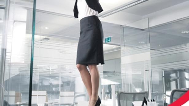 Eine Frau in Businesskleidung steht auf einem Schreibtisch und streicht die Decke mit einer Farbrolle.
