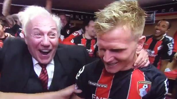 Ein älterer Mann im Anzug und ein junger Mann im Fußballtrikot jubeln gemeinsam mit anderen im Hintergrund.