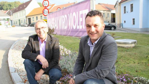 Zwei Männer hocken vor einem Banner mit der Aufschrift „Hansau wird violett“.
