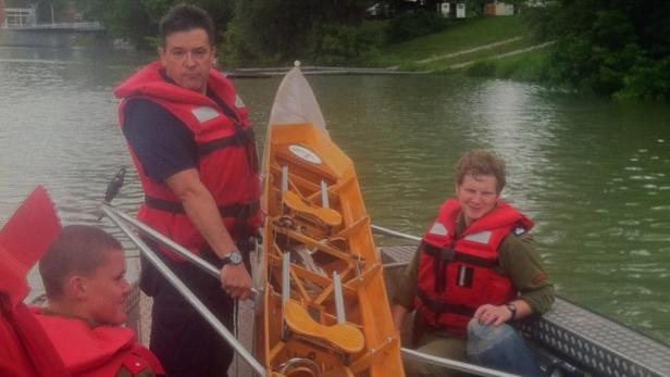 Mehrere Personen in Schwimmwesten auf einem Boot mit einem leeren Ruderboot an einem See.