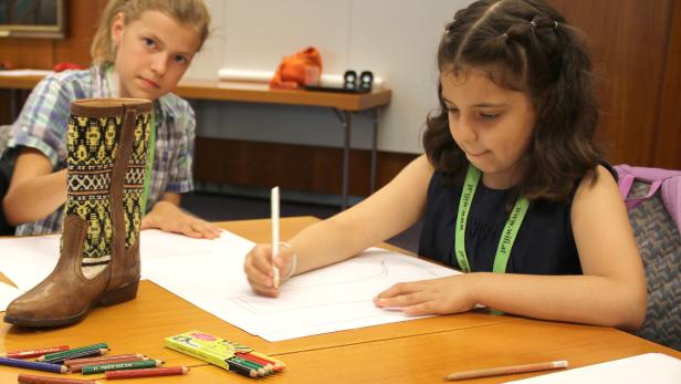 Zwei Mädchen sitzen an einem Tisch und zeichnen, ein Cowboy-Stiefel steht daneben.