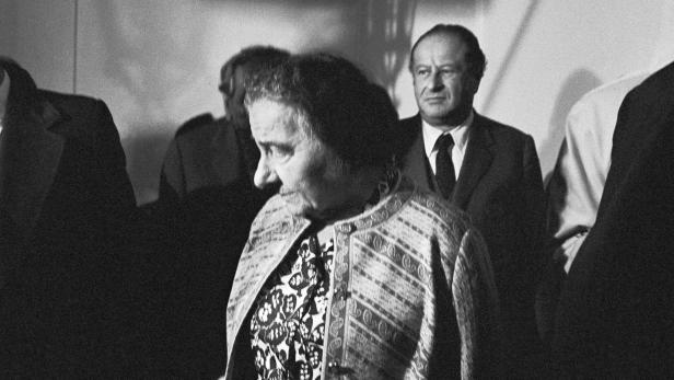 Golda Meir bei einer Veranstaltung, im Hintergrund ein Mann im Anzug.
