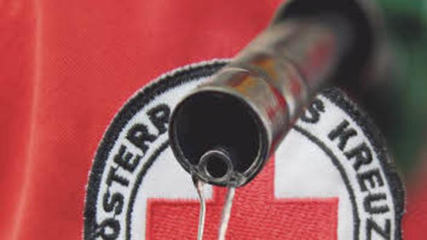 Ein Tropfen fällt aus einer Kanüle vor dem Hintergrund des Logos des Österreichischen Roten Kreuzes.