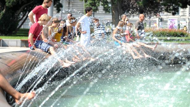 Eine Gruppe von Menschen sitzt an einem sonnigen Tag an einem Brunnen und lässt die Füße ins Wasser baumeln.