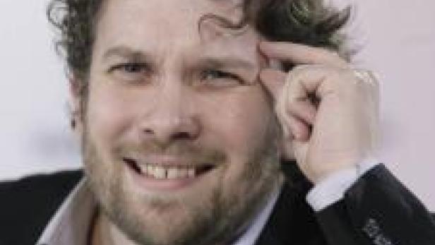 Ein Mann mit lockigem Haar lächelt und berührt seine Stirn.