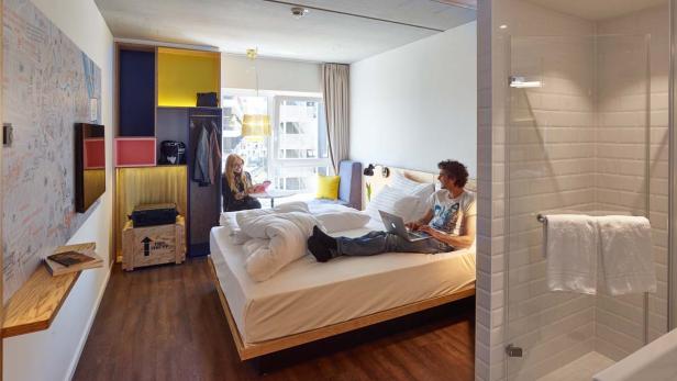 Ein Mann liegt mit einem Laptop auf dem Bett, während eine Frau am Tisch sitzt.
