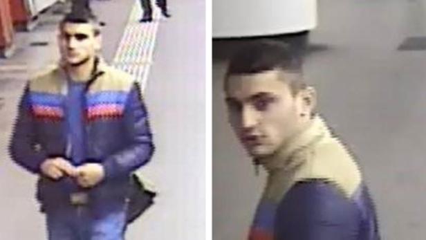 Ein junger Mann mit dunklen Haaren und einer blau-braun-roten Jacke steht auf einem Bahnsteig.