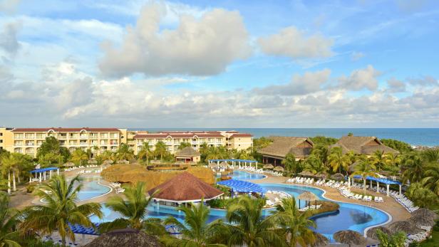 Ein Hotelresort mit Pools und Palmen am Meer unter einem bewölkten Himmel.