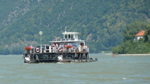 Eine Personenfähre überquert einen Fluss, im Hintergrund ein bewaldeter Hügel.