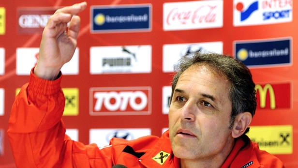 Ein Mann in roter Jacke gestikuliert vor einer Wand mit Sponsorenlogos.