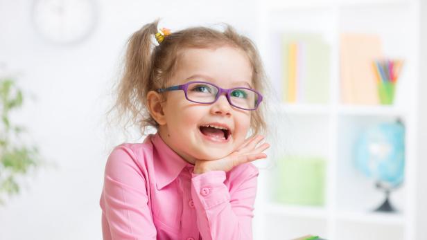 Ein lachendes Mädchen mit Brille und rosa Hemd sitzt an einem Tisch.