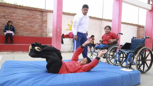 Ein Junge liegt auf einer blauen Matte, während ein Mann und ein Mädchen im Rollstuhl zusehen.