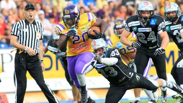Ein Footballspieler in gelb-violetter Uniform wird von einem Gegenspieler angegriffen, während ein Schiedsrichter zusieht.
