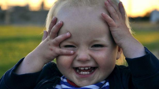 Ein lachendes Kind mit blonden Haaren hält die Hände vors Gesicht.