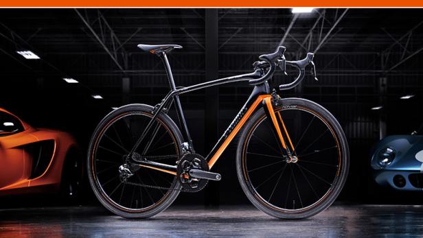 Ein Rennrad steht vor einem orangefarbenen und einem blauen Sportwagen in einer Garage.