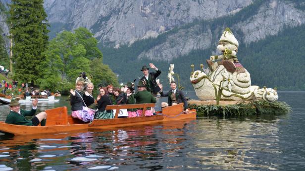 Menschen in Tracht fahren mit Booten auf einem See, im Hintergrund Berge.