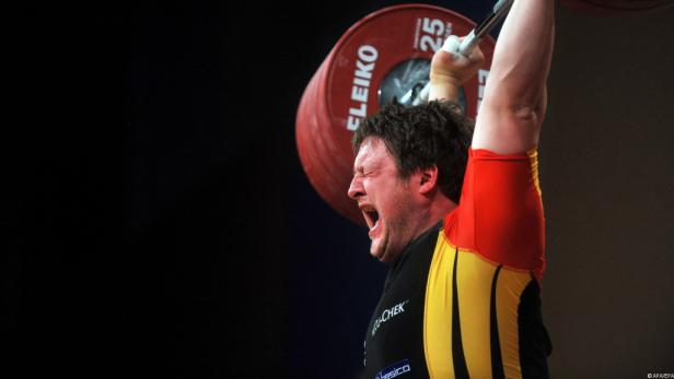 Ein Gewichtheber stemmt eine schwere Hantel mit angestrengtem Gesichtsausdruck.