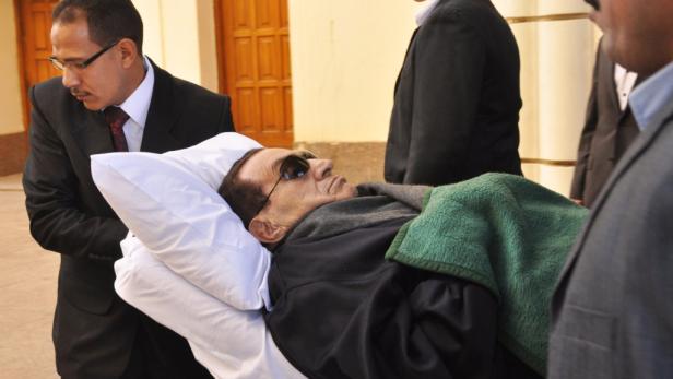 Ein Mann mit Sonnenbrille liegt auf einem Kissen, umgeben von Menschen in Anzügen.