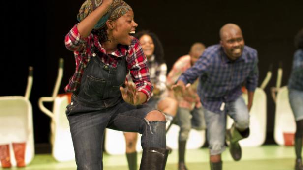 Eine Gruppe von Schauspieler:innen in Overalls und Gummistiefeln tanzt auf einer Bühne.