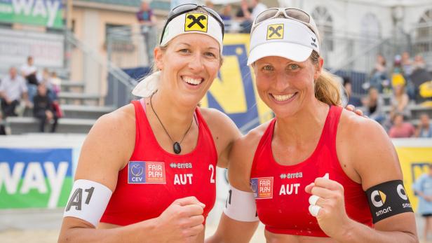 Zwei Beachvolleyballspielerinnen aus Österreich lächeln und feiern einen Sieg.