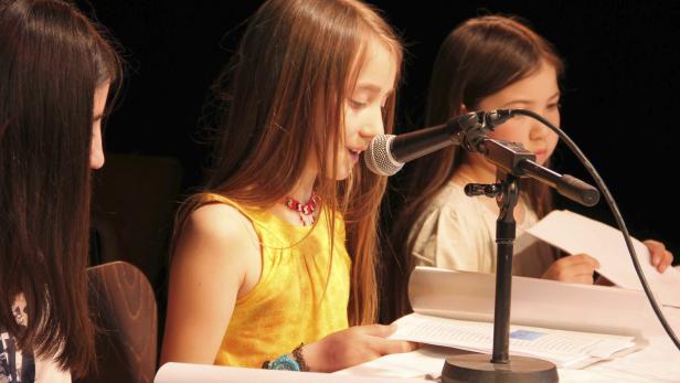 Ein junges Mädchen liest am Mikrofon vor, flankiert von zwei weiteren Mädchen.