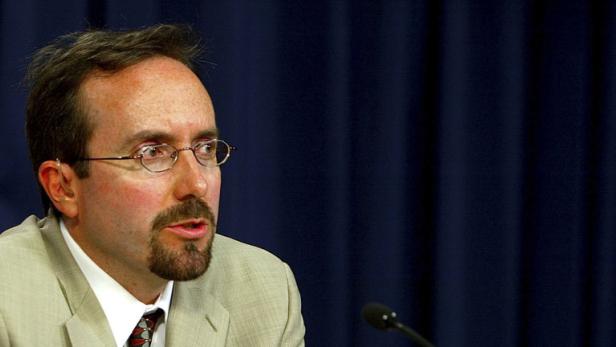 Ein Mann mit Brille und Bart spricht vor einem dunklen Hintergrund.