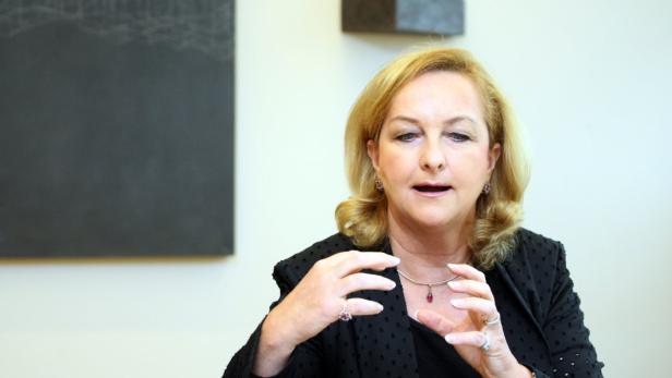 Eine Frau mit blonden Haaren gestikuliert vor einem hellen Hintergrund.