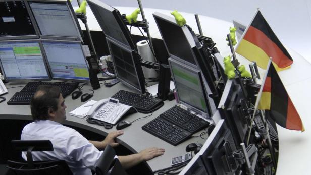 Ein Börsenhändler sitzt vor mehreren Monitoren mit Deutschland-Flaggen und kleinen grünen Figuren.