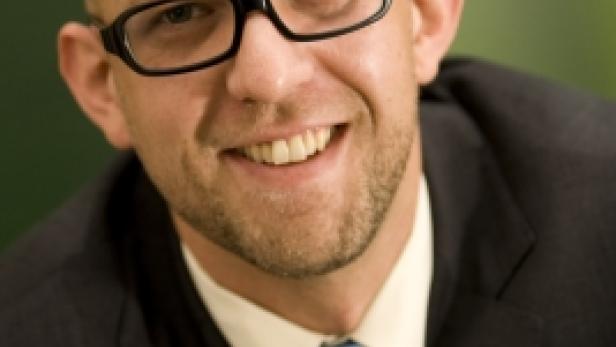 Ein lächelnder Mann mit Brille, Anzug und Krawatte vor einem grünen Hintergrund.
