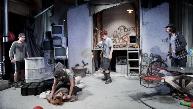 Auf einer Bühne sind mehrere junge Schauspieler in einer heruntergekommenen Umgebung zu sehen.