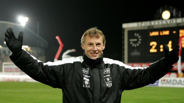 Ein lächelnder Fußballtrainer mit erhobenen Armen vor einer Anzeigetafel.