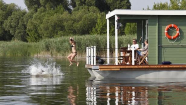 Eine Frau springt von einem Hausboot in einen See, während zwei Personen auf dem Deck sitzen.