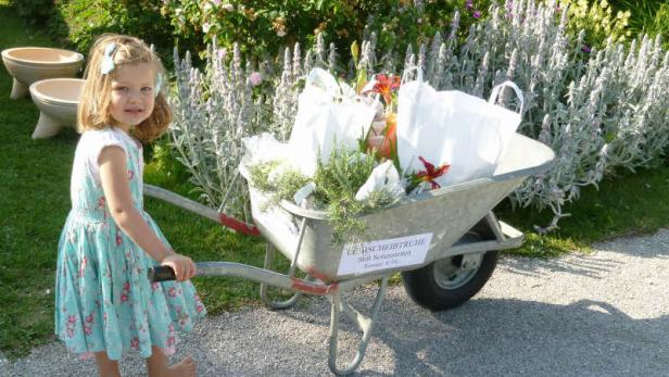 Ein kleines Mädchen schiebt barfuß eine Schubkarre voller Pflanzen und Blumen.